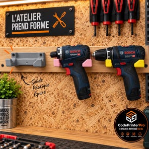 Peut inclure: Un système de rangement mural pour outils comprenant un support gris imprimé en 3D, un support rose tenant une perceuse Bosch bleue et noire, et un support jaune tenant une perceuse Bosch bleue et noire. Un panneau indique L'ATELIER PREND FORME.