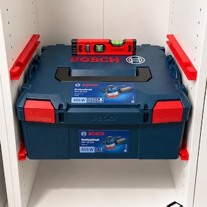 Peut inclure: Une mallette à outils Bosch L-BOXX bleu foncé avec des loquets rouges et une poignée noire, portant des étiquettes pour une ponceuse 400W Professional GEX 40-150. Un niveau à bulle Bosch rouge et noir est posé horizontalement sur le coffret.