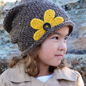 May include: A brown crocheted beanie with a yellow flower embellishment and a brown button in the centre. The beanie is worn by a child with brown hair and a tan jacket.