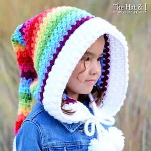 Può includere: Un cappello con cappuccio all'uncinetto arcobaleno con un bordo all'uncinetto bianco e pon pon. Il cappello è indossato da un bambino che indossa una giacca di jeans.