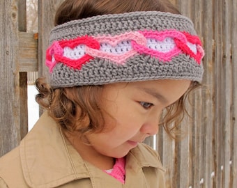 May include: A gray crocheted headband with a row of pink and white hearts. The headband is worn by a child with brown hair.