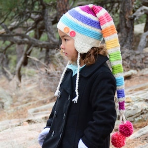 Puede incluir: Un niño lleva un gorro de crochet a rayas de colores con una cola larga y pompones. El gorro tiene orejeras y botones. El niño lleva un abrigo negro y guantes blancos.