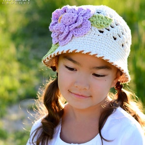 Puede incluir: Una niña pequeña con un sombrero de sol blanco de crochet con una flor morada y hojas verdes. El sombrero tiene un ribete marrón alrededor del borde. La niña lleva una camisa blanca y tiene el pelo en trenzas.