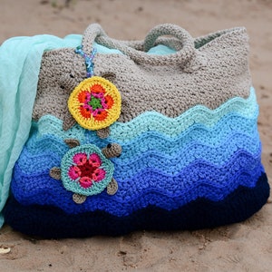 May include: A crocheted beach bag with a wavy design in shades of blue, teal, and gray. The bag features two crocheted turtle embellishments with colorful flower centers. The bag is on a sandy beach.