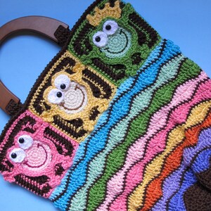 Pode incluir: Uma bolsa de mão de crochê colorida com uma alça de madeira marrom. A bolsa apresenta um padrão repetitivo de sapos com olhos de diferentes cores e um padrão de listras onduladas.