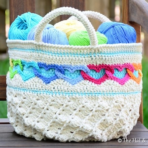 May include: A crocheted basket with a white base and colourful heart-shaped trim. The basket is filled with balls of yarn in various shades of blue, green, and yellow.