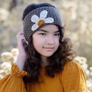 May include: A brown crocheted headband with a white crocheted flower and a yellow center. The headband is worn by a person with long brown hair and a yellow shirt.