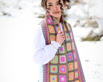 May include: A long, brown crochet scarf with colorful granny squares and fringe. The scarf is made with a variety of colors, including pink, blue, green, yellow, and orange. The scarf is worn by a person in a white shirt and blue jeans.