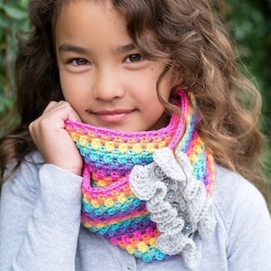 May include: A colorful rainbow crochet cowl with a gray crocheted ruffle and buttons. The cowl is worn by a young person with long brown hair.