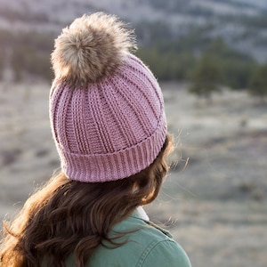 May include: A pink crocheted beanie with a brown faux fur pom pom. The beanie is worn by a person standing in a field with a mountain range in the background.