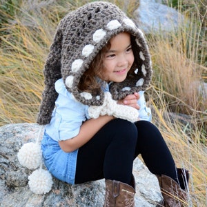 Może przedstawiać: Mała dziewczynka w brązowo-białym, dzianym kapturze z pomponami. Kaptur ma białe kropki. Dziewczynka siedzi na skale i ma na sobie brązowe kowbojskie buty.