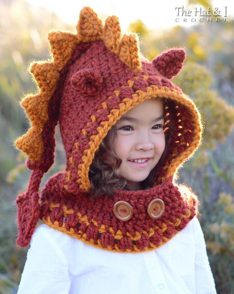 May include: A child wearing a red and orange crocheted dragon hooded cowl with wooden buttons. The cowl has a long tail and a spiky hood.