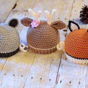 May include: Three crocheted animal hats, a raccoon, a deer, and a fox, are lined up on a wooden surface. The raccoon hat is gray with black ears, the deer hat is brown with white antlers and a pink flower, and the fox hat is orange with black ears and a black tail.