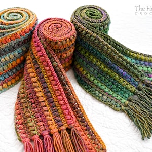 Puede incluir: Tres bufandas de crochet en diferentes combinaciones de colores, cada una con una franja en el extremo. Las bufandas est&aacute;n enrolladas y se muestran sobre una superficie blanca. El texto "The Hat & I CROCHET" es visible en la esquina superior derecha de la imagen.