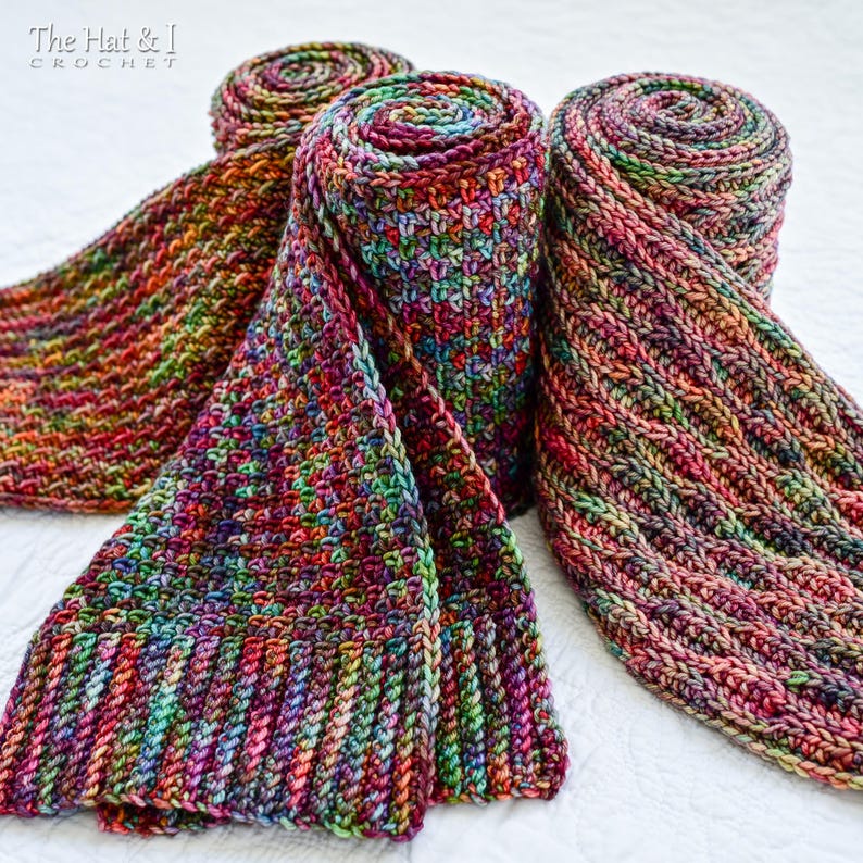 May include: Three colorful, hand-crocheted scarves in shades of red, purple, green, and orange. Two scarves are rolled up, while the third is partially unrolled, displaying its textured pattern. The scarves are made of soft yarn.