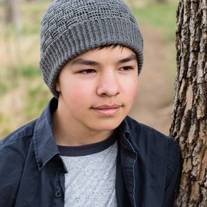 May include: A grey crocheted beanie with a textured stitch pattern. The beanie is being worn by a person with dark hair and brown eyes.