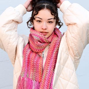 Pu&ograve; includere: Una sciarpa all'uncinetto rosa e arancione con frange. La sciarpa &egrave; indossata da una persona con i capelli ricci e una giacca trapuntata bianca.