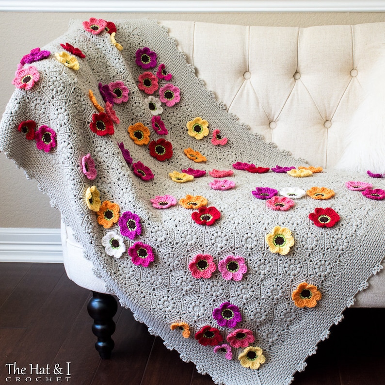 May include: A crocheted throw blanket in a light gray color with a delicate floral pattern. The blanket is adorned with numerous crocheted flowers in various shades of pink, red, orange, and yellow.