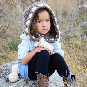 May include: A young person wearing a brown and white crocheted hooded scarf with pom poms. The scarf has a white polka dot pattern. The person is wearing a blue shirt and black pants.