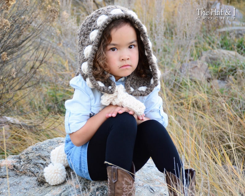 May include: A young person wearing a brown and white crocheted hooded scarf with pom poms. The scarf has a white polka dot pattern. The person is wearing a blue shirt and black pants.