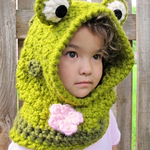 May include: A green crocheted frog hooded scarf with a pink flower detail. The scarf is worn by a child with curly brown hair.