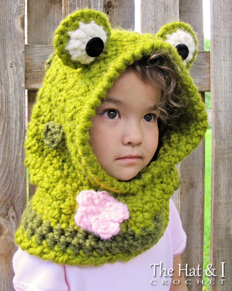 May include: A green crocheted frog hooded scarf with a pink flower detail. The scarf is worn by a child with curly brown hair.