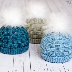 May include: Three crocheted hats with white faux fur pom poms. The hats are in shades of blue and grey.