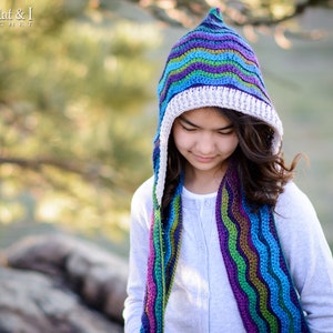 Pu&ograve; includere: Una ragazza indossa una sciarpa con cappuccio a righe colorate all'uncinetto. La sciarpa ha un bordo in crochet bianco. La sciarpa &egrave; realizzata con un motivo a punto ondulato.