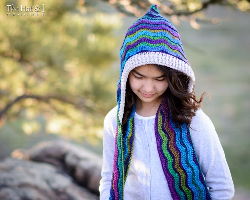 Pu&ograve; includere: Una ragazza indossa una sciarpa con cappuccio a righe colorate all'uncinetto. La sciarpa ha un bordo in crochet bianco. La sciarpa &egrave; realizzata con un motivo a punto ondulato.