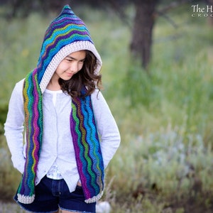 Pu&ograve; includere: Una ragazza indossa una sciarpa con cappuccio all'uncinetto colorata con un motivo ondulato. La sciarpa &egrave; realizzata con una variet&agrave; di colori, tra cui blu, verde, viola e rosa. Il cappuccio &egrave; foderato con un tessuto all'uncinetto color crema. La ragazza indossa un maglione grigio chiaro e pantaloncini di jeans blu. Lo sfondo &egrave; un'immagine sfocata di erba verde e alberi. The Hat & I Crochet &egrave; scritto nell'angolo in alto a destra dell'immagine.