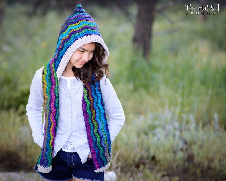 Pu&ograve; includere: Una ragazza indossa una sciarpa con cappuccio all'uncinetto colorata con un motivo ondulato. La sciarpa &egrave; realizzata con una variet&agrave; di colori, tra cui blu, verde, viola e rosa. Il cappuccio &egrave; foderato con un tessuto all'uncinetto color crema. La ragazza indossa un maglione grigio chiaro e pantaloncini di jeans blu. Lo sfondo &egrave; un'immagine sfocata di erba verde e alberi. The Hat & I Crochet &egrave; scritto nell'angolo in alto a destra dell'immagine.