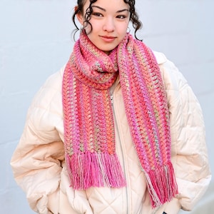 Pu&ograve; includere: Una sciarpa all'uncinetto rosa e arancione con frange. La sciarpa &egrave; indossata da una persona con i capelli castani ricci.