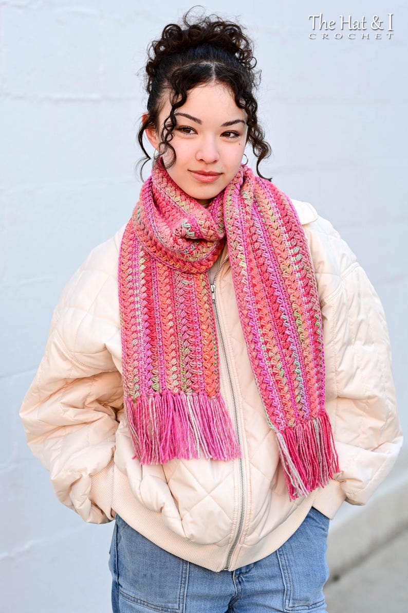 Pu&ograve; includere: Una sciarpa all'uncinetto rosa e arancione con frange. La sciarpa &egrave; indossata da una persona con i capelli castani ricci.