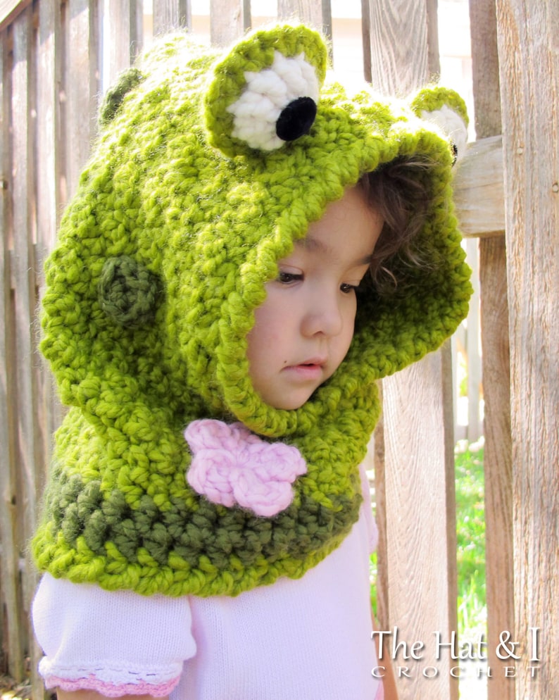 May include: Green crocheted frog hooded scarf with a pink flower detail. The hat has black eyes and white accents. The Hat & I Crochet.