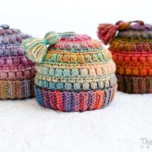 May include: Three colorful crocheted hats with tassels. The hats are made with a variety of colors, including pink, blue, green, and orange. The hats are sitting on a white surface.