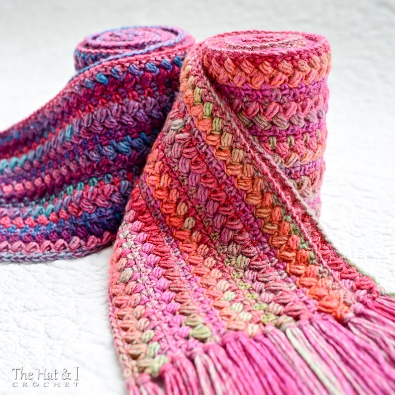 Pu&ograve; includere: Due sciarpe all'uncinetto in tonalit&agrave; rosa, viola e verde con frange. Le sciarpe sono arrotolate e mostrate su una superficie bianca. Il testo "The Hat & I Crochet" &egrave; visibile nell'angolo in basso a sinistra dell'immagine.