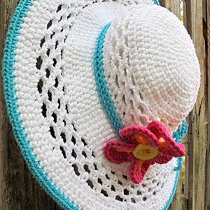Puede incluir: Sombrero de sol de crochet blanco con ribete turquesa y una flor rosa con un botón amarillo en el centro.