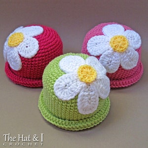 May include: Three crocheted hats with flower embellishments. The hats are in pink, green, and white. The flowers are white with yellow centres.