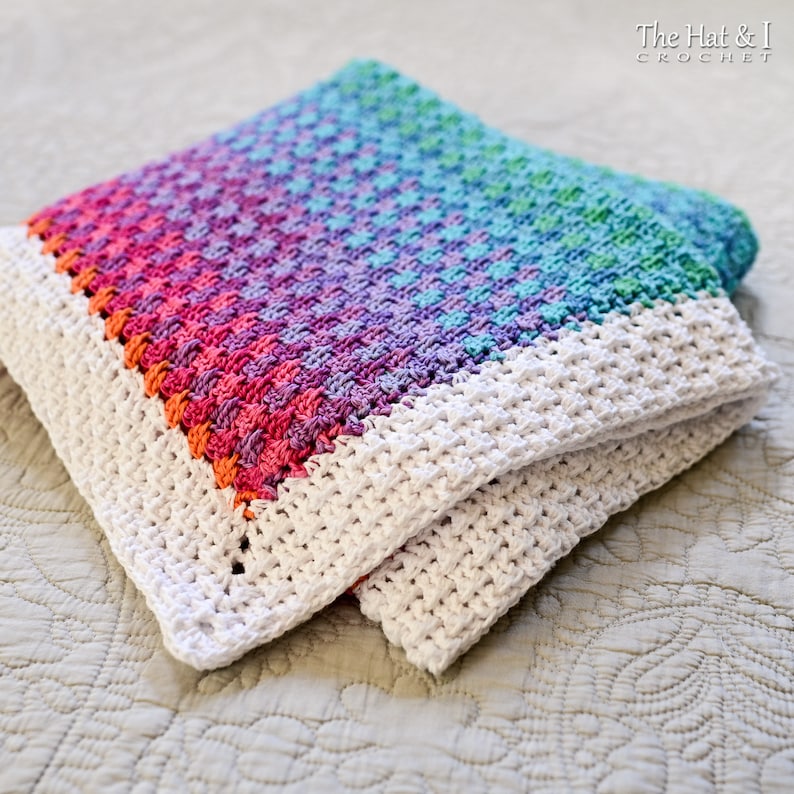 May include: A colorful crochet blanket with a rainbow ombre pattern and a white border. The blanket is folded in half and lying on a white surface.