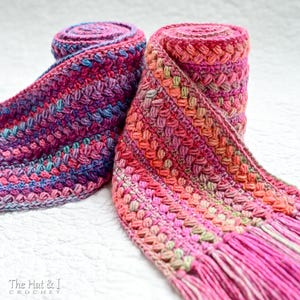 Pu&ograve; includere: Due sciarpe all'uncinetto in uno schema di colori rosa, viola e verde. Le sciarpe sono arrotolate e hanno frange alle estremit&agrave;. Le sciarpe sono su una superficie bianca. Il testo "The Hat & I Crochet" &egrave; visibile nell'angolo in basso a sinistra dell'immagine.