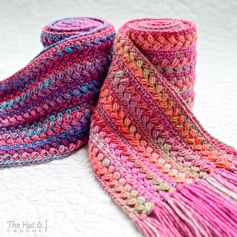 Pu&ograve; includere: Due sciarpe all'uncinetto in uno schema di colori rosa, viola e verde. Le sciarpe sono arrotolate e hanno frange alle estremit&agrave;. Le sciarpe sono su una superficie bianca. Il testo "The Hat & I Crochet" &egrave; visibile nell'angolo in basso a sinistra dell'immagine.