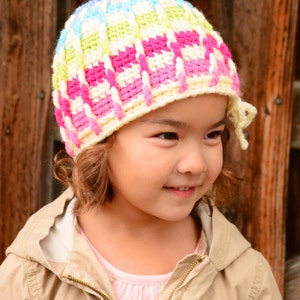 Pu&ograve; includere: Una persona giovane indossa un cappello all'uncinetto colorato con sfondo bianco e strisce rosa, blu e verdi. Il cappello ha una lunga cravatta che pende dal lato del viso della persona.