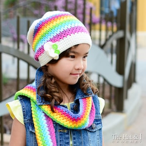 May include: A crocheted rainbow hat and scarf set. The hat is white with a rainbow stripe design and a white flower with green button accents. The scarf is a matching rainbow color. The Hat & I Crochet logo is in the bottom right corner.