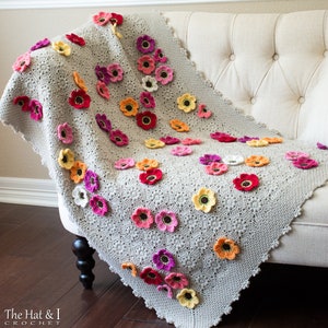 May include: A crocheted throw blanket with a gray background and a repeating pattern of colorful flowers. The blanket is draped over a white couch with tufted upholstery.