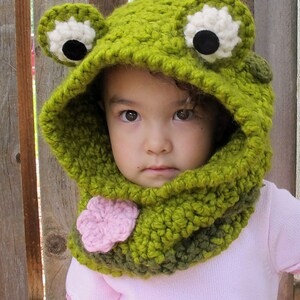 May include: A green crocheted frog hat with black eyes and a pink flower on the neck. The hat has a hood and a scarf attached. The hat is worn by a child.