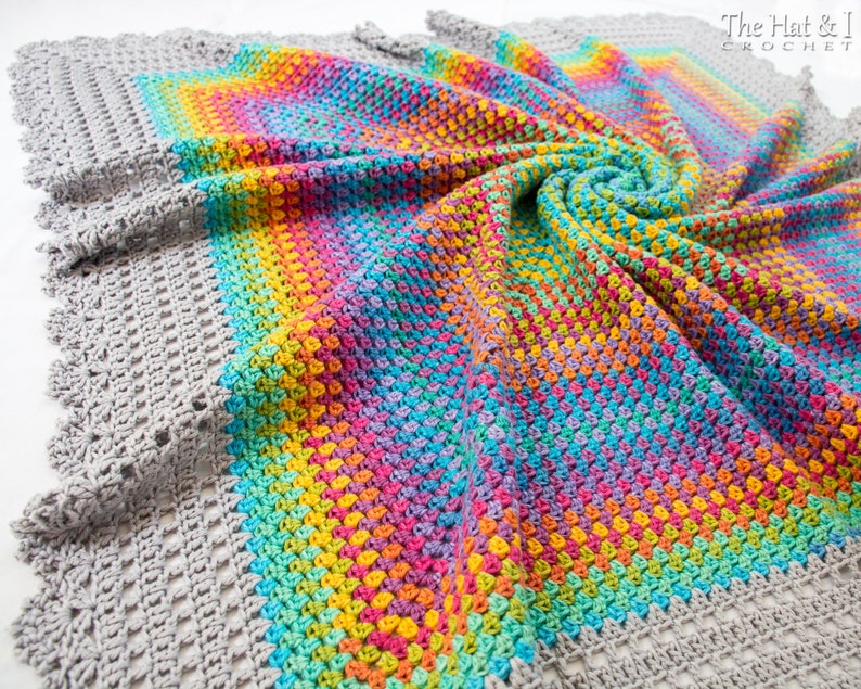 May include: A crocheted blanket with a rainbow pattern in the center and a gray crocheted border. The blanket is made with a variety of colors including pink, blue, green, yellow, and orange. The blanket is folded in a spiral shape. The text 'The Hat & I CROCHET' is visible in the upper right corner of the image.