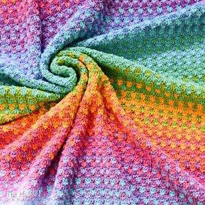 May include: A colorful crochet blanket with a rainbow pattern. The blanket is made with a textured stitch and is folded in a spiral shape.