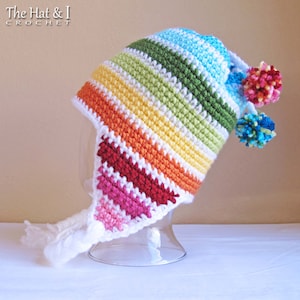 May include: A rainbow striped crochet hat with a white brim and pom poms. The hat has a long white tassel hanging from the brim. The hat is on a white surface. The Hat & I CROCHET is written in the top left corner of the image.