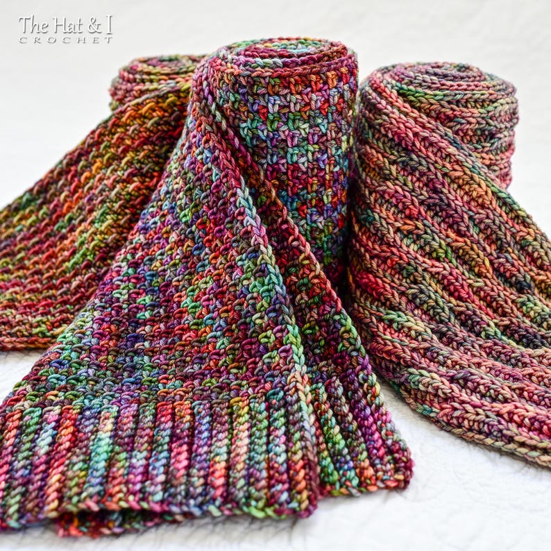 May include: Three colorful, hand-crocheted scarves in shades of red, purple, green, and orange. Two scarves are rolled up, while the third is partially unfolded, displaying the intricate stitch pattern. The scarves are made of a soft, textured yarn.