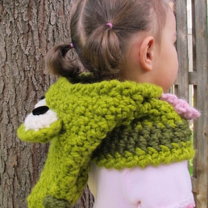 May include: A green and brown crocheted hooded scarf with a frog face on the hood. The scarf is worn by a child with pigtails.